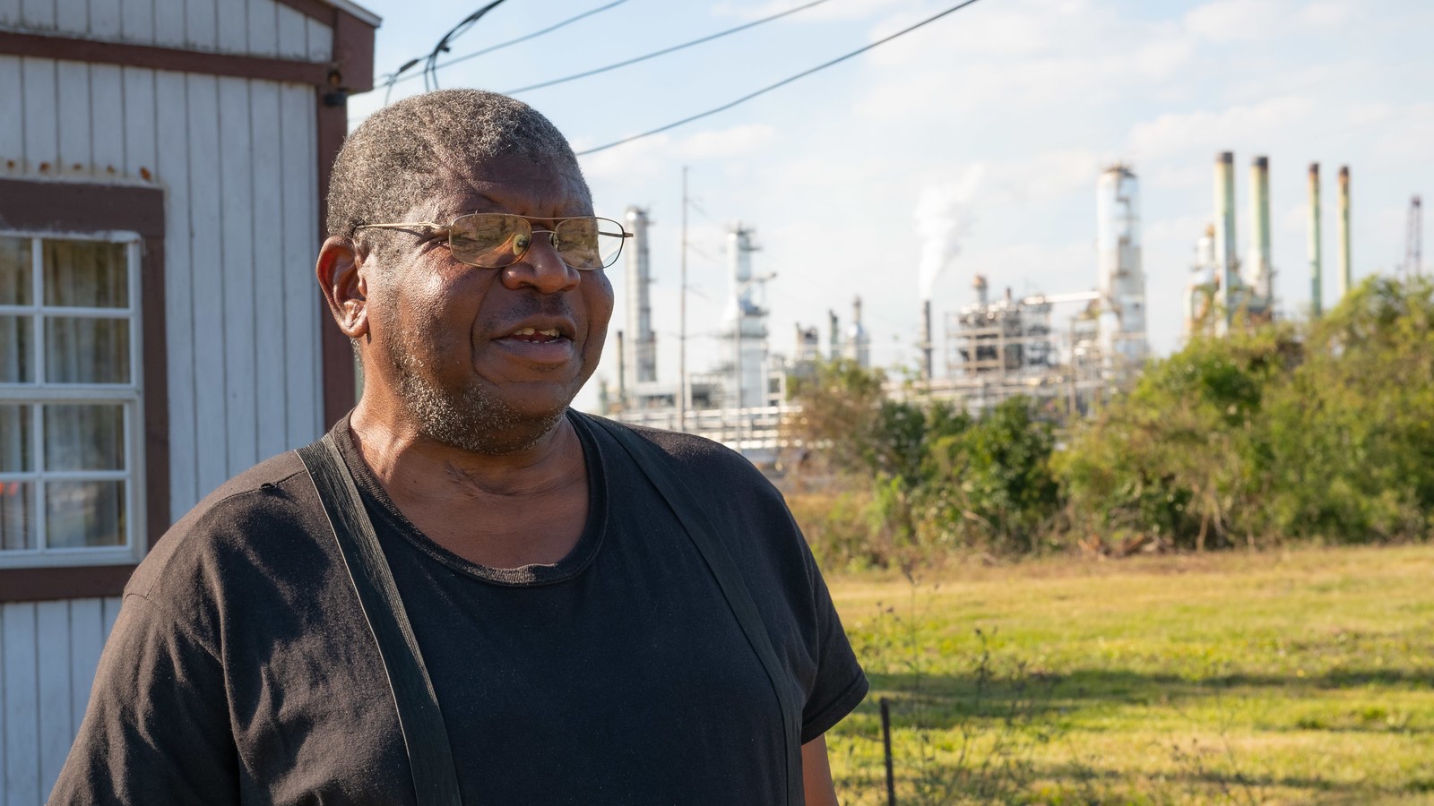 Black man with glasses and grey hair. A fossil fuel refinery can be seen behind him in the distance.