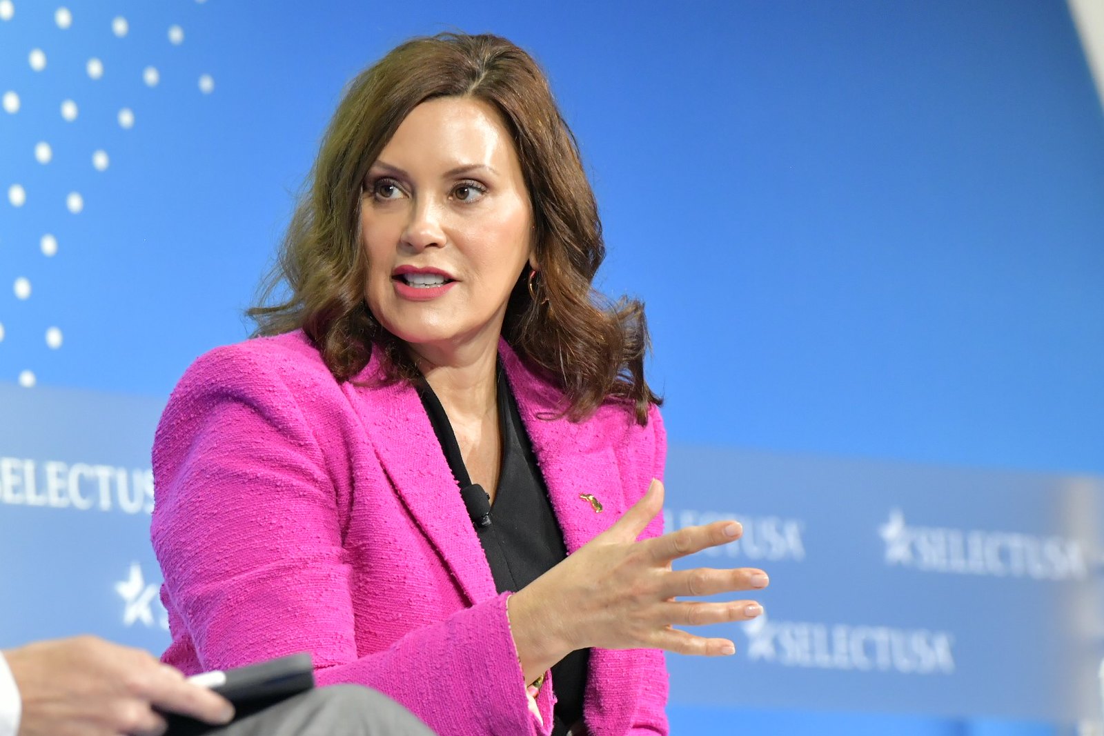 Michigan Governor Gretchen Whitmer speaks while seated on a stage with the Select USA logo in the background