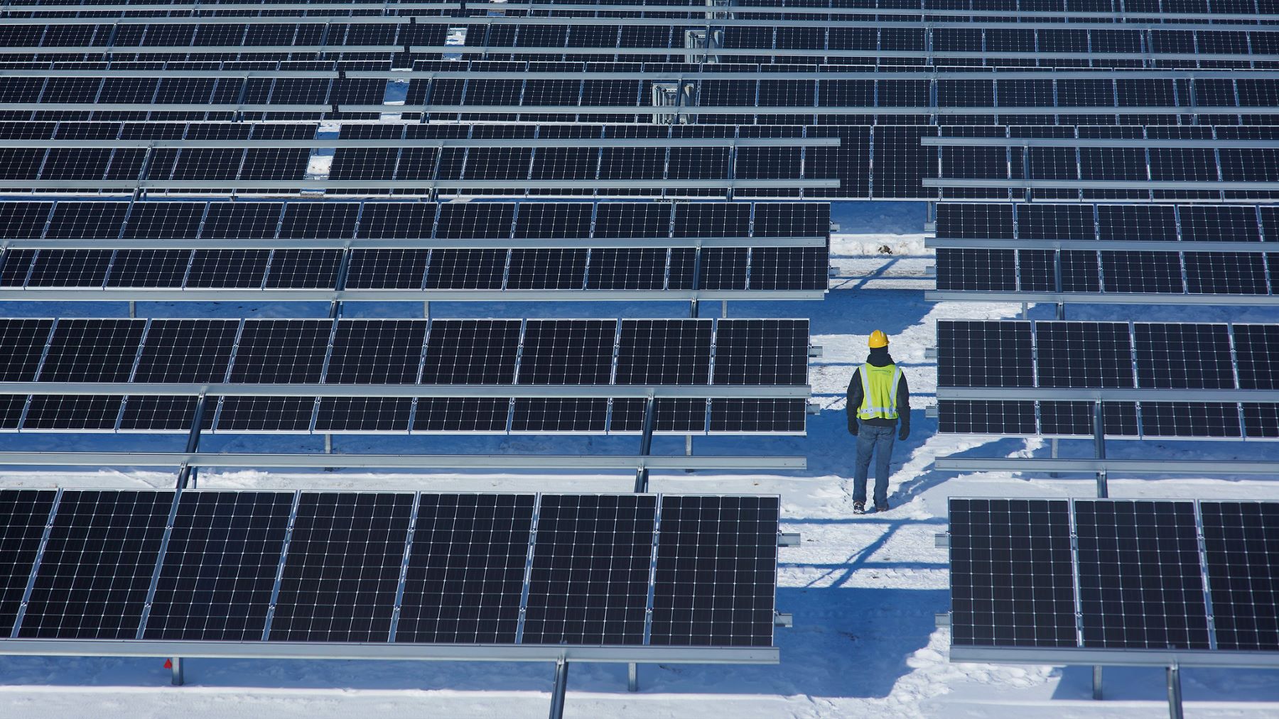 Wide shot of a solar panel farm on a snowy day. A worker with a yellow hard hat and neon yellow vest stands amongst the panels, with their back towards the camera.