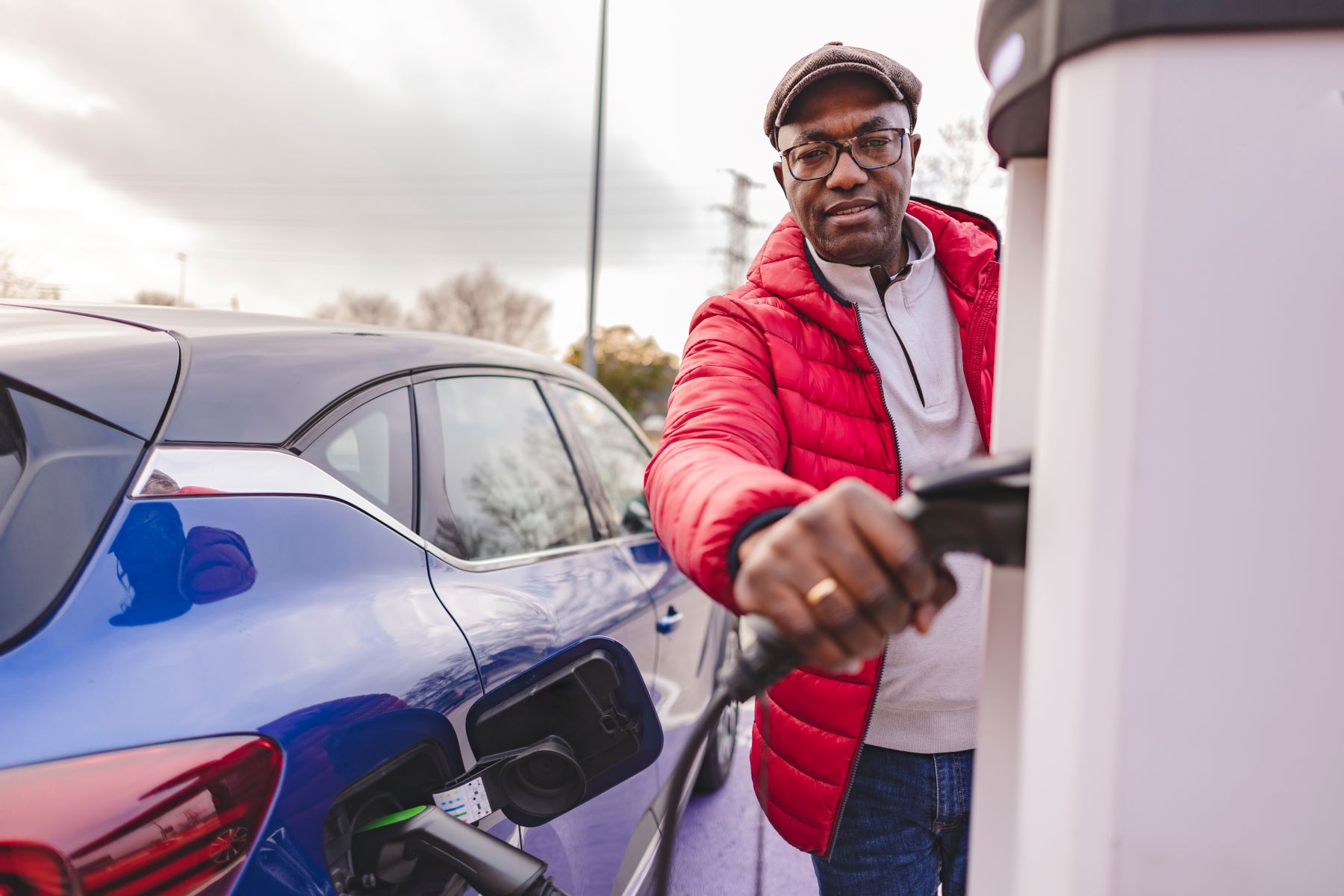 Black man with red jacket and brown hat grabbing an electric car charger to plug into his blue EV.