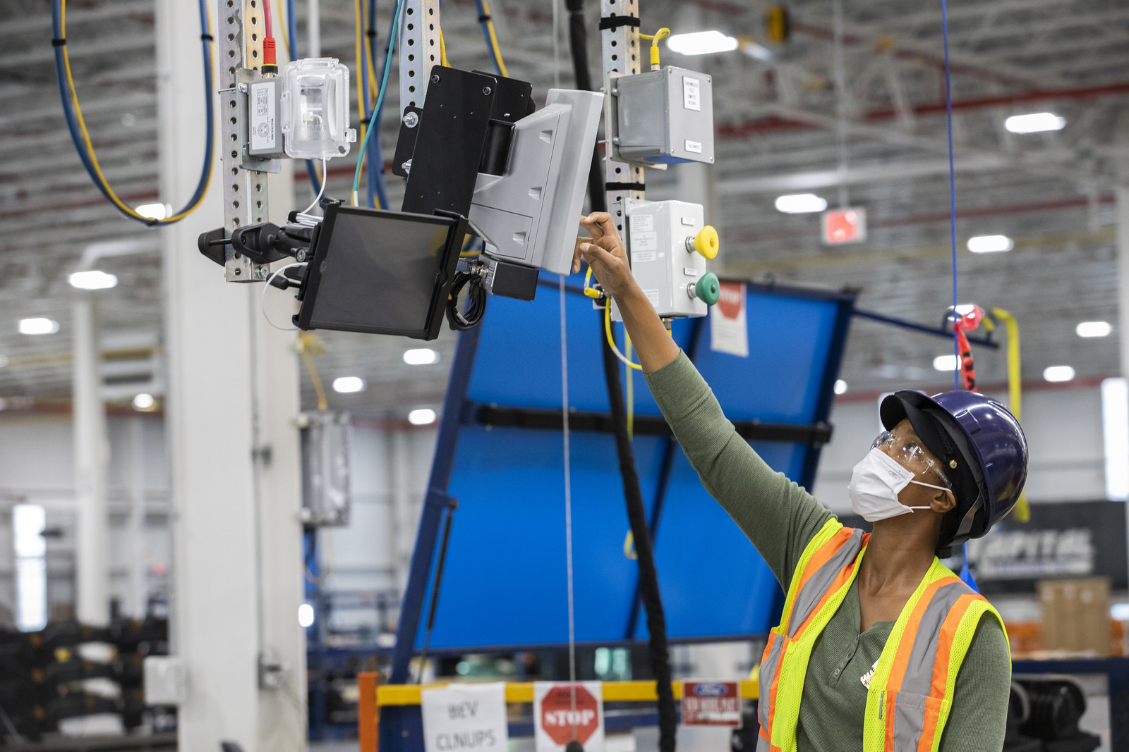 A woman in safety equipment reaches for a screen above her in an electric vehicle manufacturing facility.