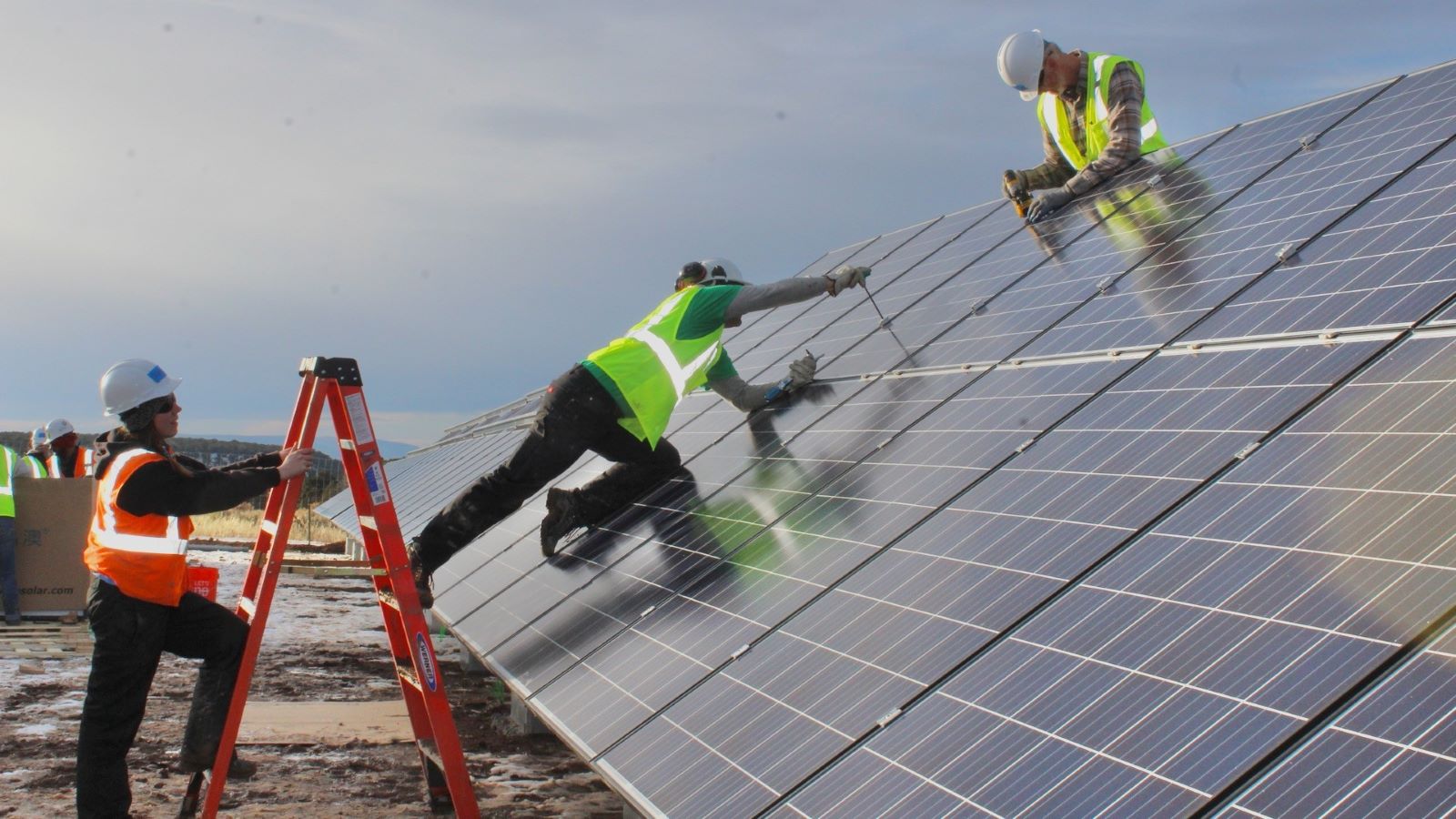 3 nonprofit solar installers and volunteers building a solar array in Colorado. The worker on the left is on a ladder, one is on the panel screwing a piece in, and another at the top is drilling.