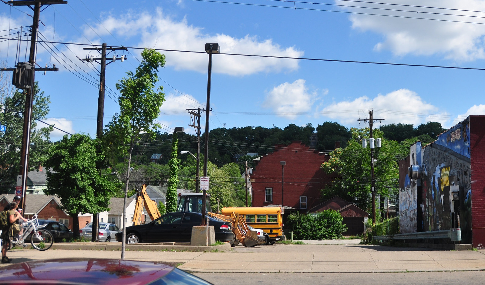 Wide shot of a bustling street in Philadelphia, Pennsylvania. People are walking and biking. School buses are parked on the side of the street. A mural is seen in the back. 