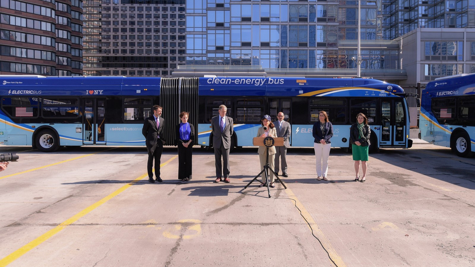 Wide shot of New York Governor Hochul speaking at a podium in front of a blue electric bus. Behind her are other government officials. 